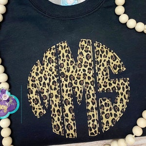 May include: Black sweatshirt with a large, circular monogram in a leopard print design. The monogram is made up of the letters "MME".