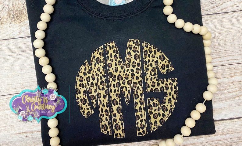May include: Black sweatshirt with a large, circular monogram in a leopard print design. The monogram is made up of the letters "MME".