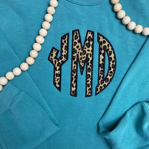 May include: A teal blue sweatshirt with a large, round, black-outlined monogram in a leopard print pattern. The monogram is "YMD".