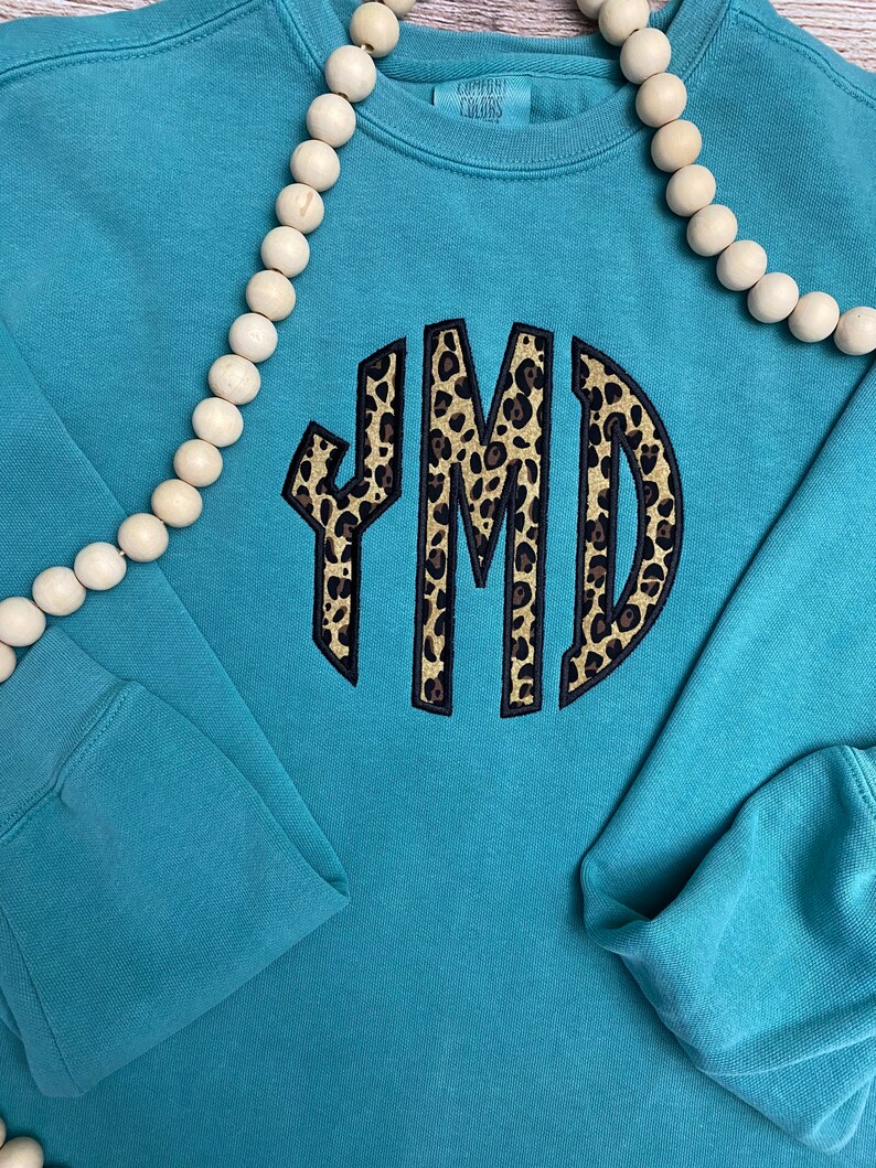 May include: A teal blue sweatshirt with a large, round, black-outlined monogram in a leopard print pattern. The monogram is "YMD".