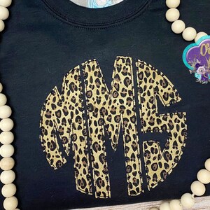 May include: Black t-shirt with a large, circular monogram in a leopard print pattern. The monogram is made up of the letters "MME".