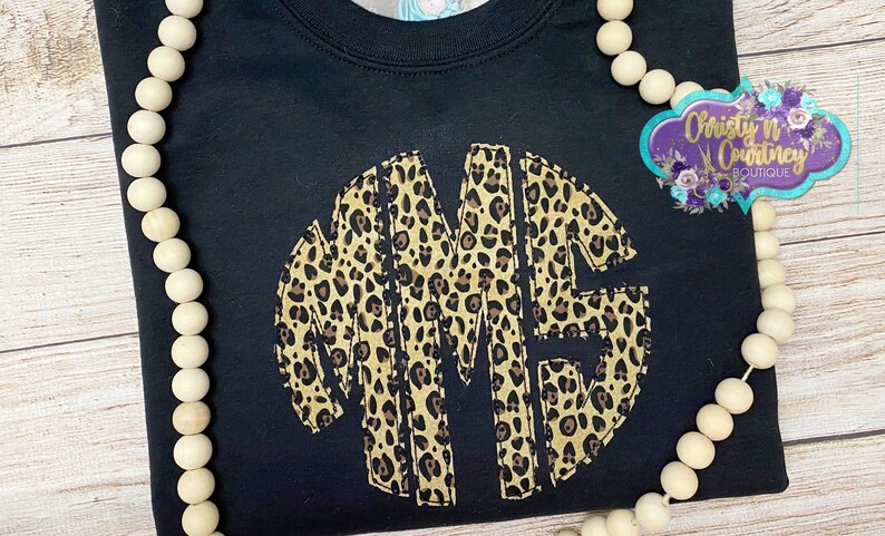 May include: Black t-shirt with a large, circular monogram in a leopard print pattern. The monogram is made up of the letters "MME".
