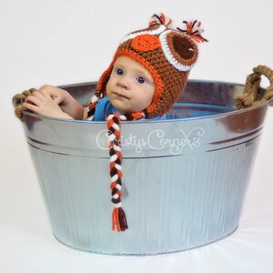 K&ouml;nnte beinhalten: Ein Baby tr&auml;gt eine orangefarbene, braune und wei&szlig;e geh&auml;kelte Eulenm&uuml;tze und sitzt in einer silbernen Metallwanne. Die M&uuml;tze hat einen langen, geflochtenen Schwanz.