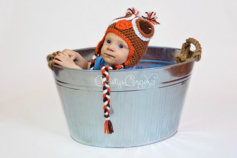 K&ouml;nnte beinhalten: Ein Baby tr&auml;gt eine orangefarbene, braune und wei&szlig;e geh&auml;kelte Eulenm&uuml;tze und sitzt in einer silbernen Metallwanne. Die M&uuml;tze hat einen langen, geflochtenen Schwanz.