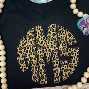 May include: Black t-shirt with a large, circular monogram in a leopard print pattern. The monogram is made up of the letters "MME".