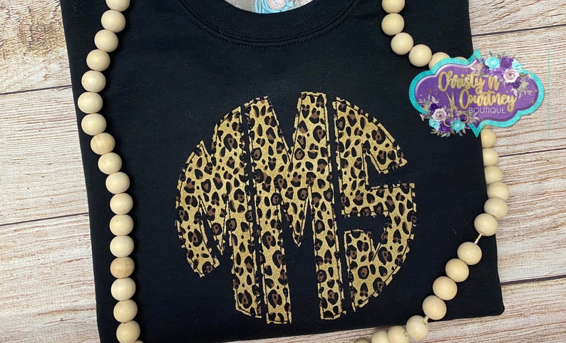 May include: Black t-shirt with a large, circular monogram in a leopard print pattern. The monogram is made up of the letters "MME".