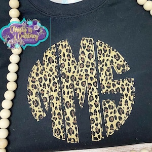 May include: Black sweatshirt with a large, circular, leopard print monogram. The monogram is made up of the letters "M" and "E".