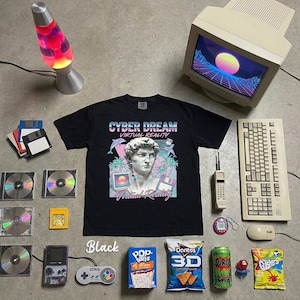 Może przedstawiać: Czarny t-shirt z napisem "CYBER DREAM VIRTUAL REALITY" i grafiką statuy, otoczony przedmiotami retro. Zawiera monitor komputera, klawiaturę, lampę lawową, dyskietki, płyty CD i Game Boy.