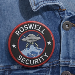 Può includere: Una toppa rotonda con le parole "ROSWELL SECURITY" che circondano un disegno di UFO blu e grigio. La toppa è nera con un bordo rosso e due stelle bianche. È cucita su una giacca di jeans blu.
