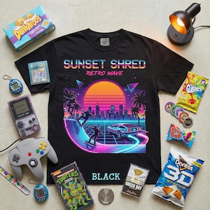 Puede incluir: Camiseta negra con diseño "SUNSET SHRED RETRO WAVE" que muestra skaters, un coche y una puesta de sol. Rodeada de objetos retro como una Game Boy, snacks y un mando de Nintendo 64.