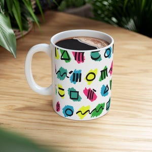 Op de afbeelding: Witte keramische koffiemok met een kleurrijk geometrisch patroon van vormen en lijnen in groen, blauw, roze en geel. De mok is gevuld met donkere koffie en staat op een houten oppervlak.