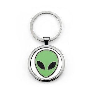Könnte beinhalten: Ein silberner Schlüsselanhänger mit einem kreisförmigen Anhänger, der ein grünes Alien-Gesicht mit schwarzen Augen zeigt. Das Alien-Design ist von einem silbernen Rahmen umgeben. Der Schlüsselanhänger hat einen Schlüsselring.
