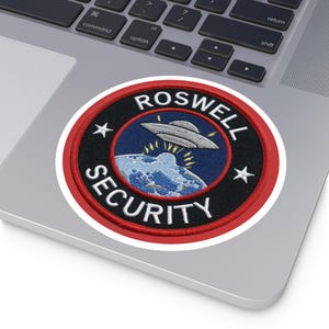 Op de afbeelding: Een ronde patch met de woorden "ROSWELL SECURITY" in witte letters. De patch toont een blauwe en witte afbeelding van een UFO boven een planeet. De patch heeft een zwarte achtergrond met een rode rand en witte sterren.