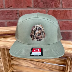 May include: Olive green flat bill snapback hat with a mesh back and a detailed embroidered dog face on the front. The hat has a Richardson tag on the front.