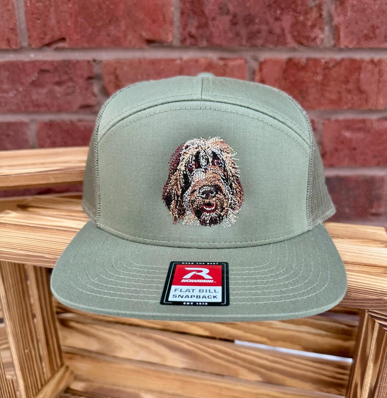May include: Olive green flat bill snapback hat with a mesh back and a detailed embroidered dog face on the front. The hat has a Richardson tag on the front.