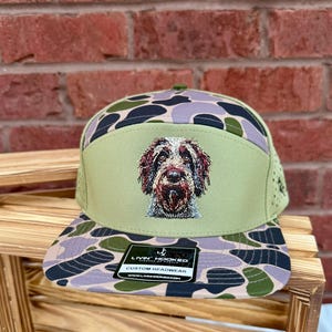 May include: A green and camouflage-patterned baseball cap featuring an embroidered image of a dog. The cap has a flat brim and a mesh back. The front panel displays the dog's face, and the cap includes a tag that reads "LIVIN' HOOKED CUSTOM HEADWEAR".