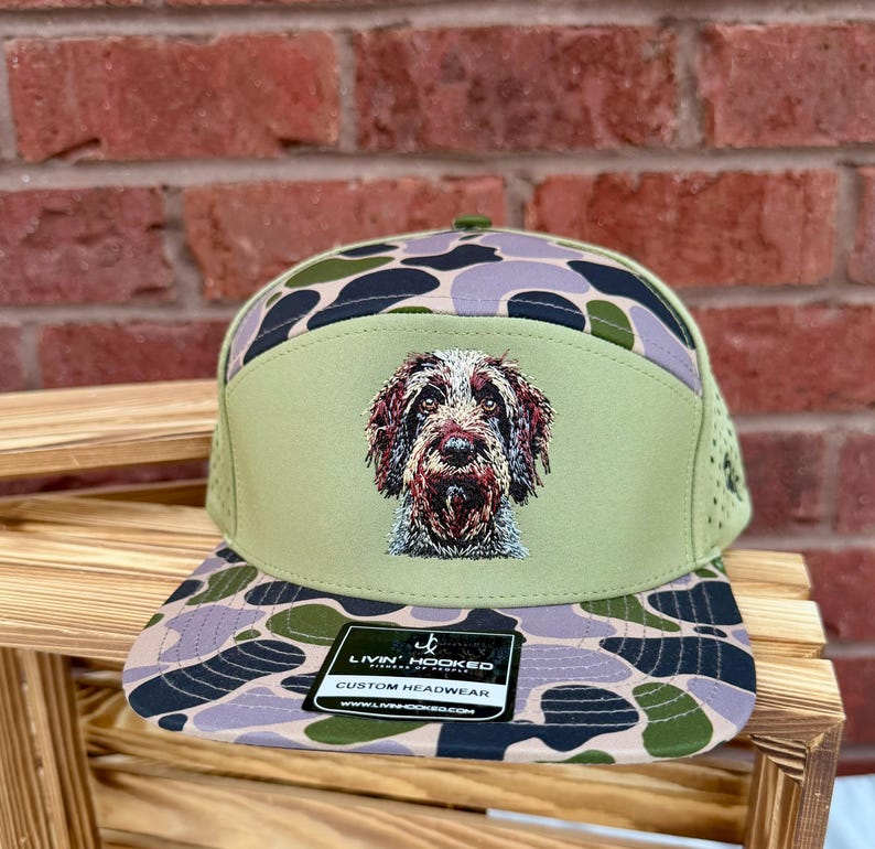 May include: A green and camouflage-patterned baseball cap featuring an embroidered image of a dog. The cap has a flat brim and a mesh back. The front panel displays the dog's face, and the cap includes a tag that reads "LIVIN' HOOKED CUSTOM HEADWEAR".