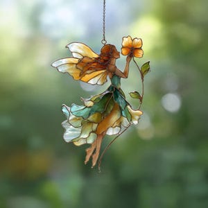 以下が含まれることがあります： 黄色と白の羽を持つステンドグラスの妖精が、黄色の花を持っています。妖精のドレスは緑、黄色、白のミックスです。妖精はチェーンで吊り下げられており、背景はぼやけた緑色です。