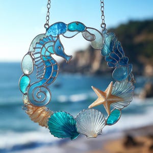 Puede incluir: Una corona de vidrieras con un caballito de mar y conchas marinas en tonos azules, turquesas y beige. El caballito de mar es el punto focal, rodeado de conchas y una estrella de mar. La corona está suspendida por una cadena, con un fondo borroso del océano.