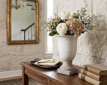 May include: A white ceramic urn-style vase with a pedestal base, filled with a bouquet of white ranunculus and tan hydrangea flowers. The vase sits on a wooden console table next to a stack of three vintage books and a black tray with a magnifying glass.