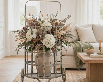 May include: A weathered, gray metal rolling cart with a cage-like structure holds a large, beige ceramic vase filled with a dried floral arrangement. The bouquet features white hydrangea blossoms, tall pampas grass, and various dried greenery.