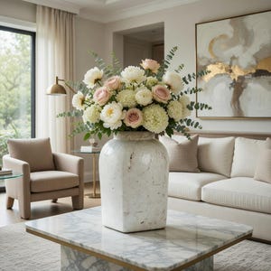 May include: A large white vase filled with a bouquet of white and pink flowers sits on a marble coffee table. The flowers include roses and peonies, with green foliage. The vase has a textured, aged appearance.