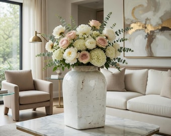May include: A large white vase filled with a bouquet of white and pink flowers sits on a marble coffee table. The flowers include roses and peonies, with green foliage. The vase has a textured, aged appearance.
