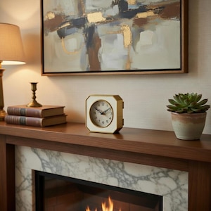 May include: A white and gold-framed clock with a cream-colored face sits on a wooden mantel. The clock has black hands and numerals. A succulent plant in a small pot is to the right of the clock. A painting hangs above the mantel.