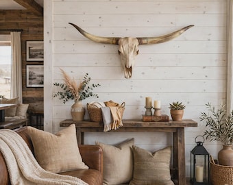 May include: A large longhorn steer skull mounted on a white horizontal plank wall above a rustic wooden console table. The table holds a woven basket with textiles, two pillar candles, a small potted succulent, and a ceramic vase with dried foliage.