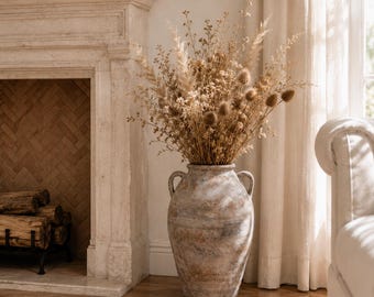 May include: A large, weathered, light brown ceramic amphora vase with two side handles and horizontal textured bands, filled with a tall, dense arrangement of dried beige grasses, feathery pampas plumes, and round, prickly brown seed heads.