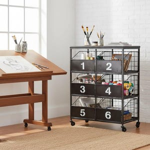 May include: A black metal storage unit with six numbered bins, ideal for an art studio or craft room. The unit is on wheels and has a wire mesh design. A wooden drafting table and art supplies are also visible.