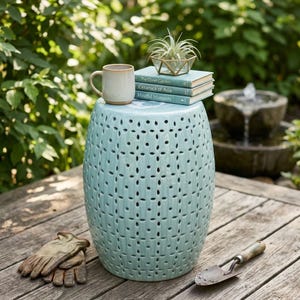 May include: A light blue ceramic garden stool with a repeating cut-out pattern, topped with a beige mug, three stacked books titled The Quiet Garden, Ceramics of Asia, and Mindful Moments, and a small air plant in a geometric glass container.