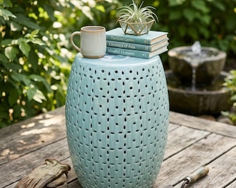 May include: A light blue ceramic garden stool with a repeating cut-out pattern, topped with a beige mug, three stacked books titled The Quiet Garden, Ceramics of Asia, and Mindful Moments, and a small air plant in a geometric glass container.