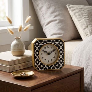 May include: A square analog clock with a gold frame and a black and white geometric pattern on the face. The clock features black numbers and hands. Beside it sits a small ceramic vase with dried grasses, two stacked books, and a gold ring dish.