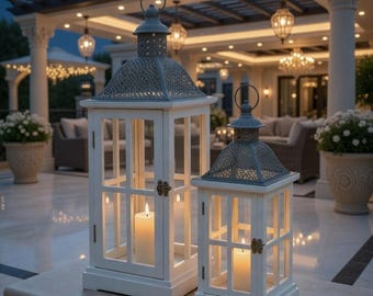 May include: Two white wooden lanterns with gray metal tops and glass panels. Each lantern has a lit candle inside, casting a warm glow. The lanterns are on a stone surface, with a blurred outdoor setting in the background.