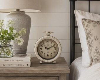May include: A round, off-white distressed table clock with a small bird figurine on top, featuring black Arabic numerals and ornate hands on a cream-colored face. The clock sits on a wooden nightstand next to a stack of books and a lamp.