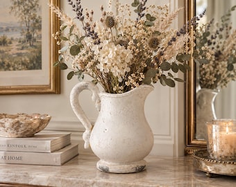 May include: A white ceramic pitcher with a distressed, weathered finish and a curved handle, filled with a dried floral arrangement featuring lavender, eucalyptus, pampas grass, and thistle, creating a neutral-toned botanical display.