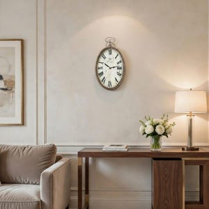 May include: Oval wall clock with Roman numerals and a distressed brown frame. The clock has a white face and black hands. The clock is hanging on a light beige wall. The clock is part of a home decor theme.
