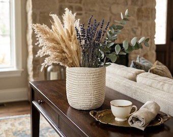 May include: A cream-colored textured ceramic vase filled with dried pampas grass, purple lavender sprigs, and green eucalyptus leaves. Next to the vase sits a white teacup with a gold rim on a small brass tray with a folded beige linen napkin.