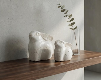 May include: Two white ceramic bunny figurines with textured ears and speckled details sit on a wooden shelf. A clear glass vase with a sprig of greenery is placed behind the bunnies, creating a minimalist, springtime aesthetic.