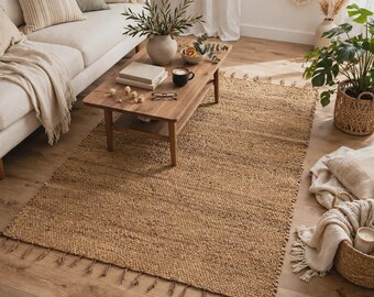 May include: A rectangular area rug crafted from natural woven jute fibers, featuring a textured, tan-colored surface and decorative fringe detailing along the short edges. The rug is positioned on a light-toned wooden floor in a living space.