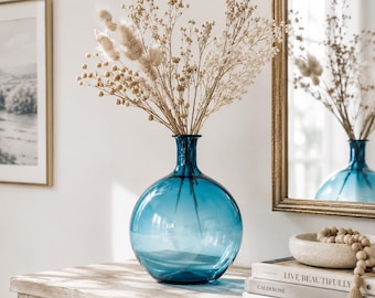 May include: A round, translucent blue glass vase with a narrow neck, containing a bouquet of dried beige grasses and fluffy stems. The vase sits on a wooden surface next to a stack of books with the text Live Beautifully, Calderone, This Is Home.