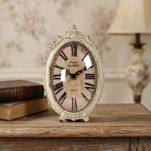 May include: An antique-style oval clock with a distressed ivory finish. The clock face features Roman numerals and the text "Fabrique de PARIS 1887". The clock is set on a wooden surface, with two antique books in the background.