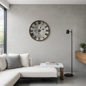 May include: A large, round wall clock with a silver face and black Roman numerals. The clock has a brown wooden frame and is mounted on a gray wall. The clock is a decorative piece for a living room or bedroom.
