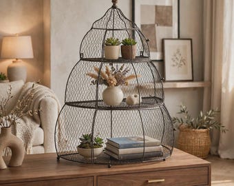 May include: A three-tiered, dome-shaped wire display rack with a dark metal finish. The shelves hold two small potted succulents, a ceramic vase with dried botanicals, a small stone-like object, a potted plant, and three stacked books.