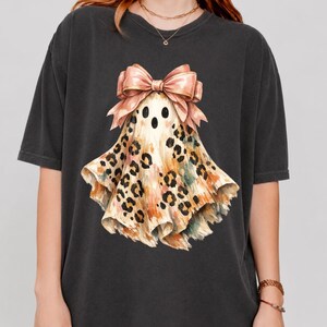 May include: A charcoal gray short-sleeve t-shirt featuring a central graphic of a ghost with a tan and brown leopard print pattern. The ghost is adorned with a large, light pink bow at the top, positioned above its black eyes and open mouth.