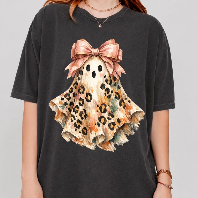 May include: A charcoal gray short-sleeve t-shirt featuring a central graphic of a ghost with a tan and brown leopard print pattern. The ghost is adorned with a large, light pink bow at the top, positioned above its black eyes and open mouth.