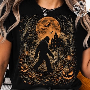May include: A black t-shirt featuring a graphic print of a large, dark, hairy creature walking through a graveyard at night. The scene includes a full orange moon, bats, a haunted house, and several glowing jack-o'-lanterns in the foreground.