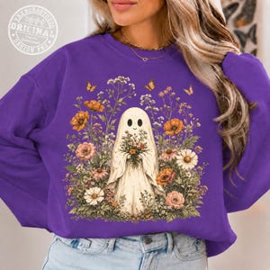 Pu&ograve; includere: Una felpa girocollo viola con una grafica centrale raffigurante un fantasma bianco che tiene in mano un bouquet di fiori, circondato da un giardino colorato di fiori selvatici arancioni, rosa e bianchi, fogliame verde e piccole farfalle gialle.