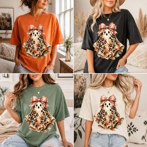 May include: A short-sleeve t-shirt available in orange, black, olive green, or cream, featuring a central graphic of a ghost with a leopard print body and a large pink bow on its head, designed with a relaxed fit and a crew neckline.